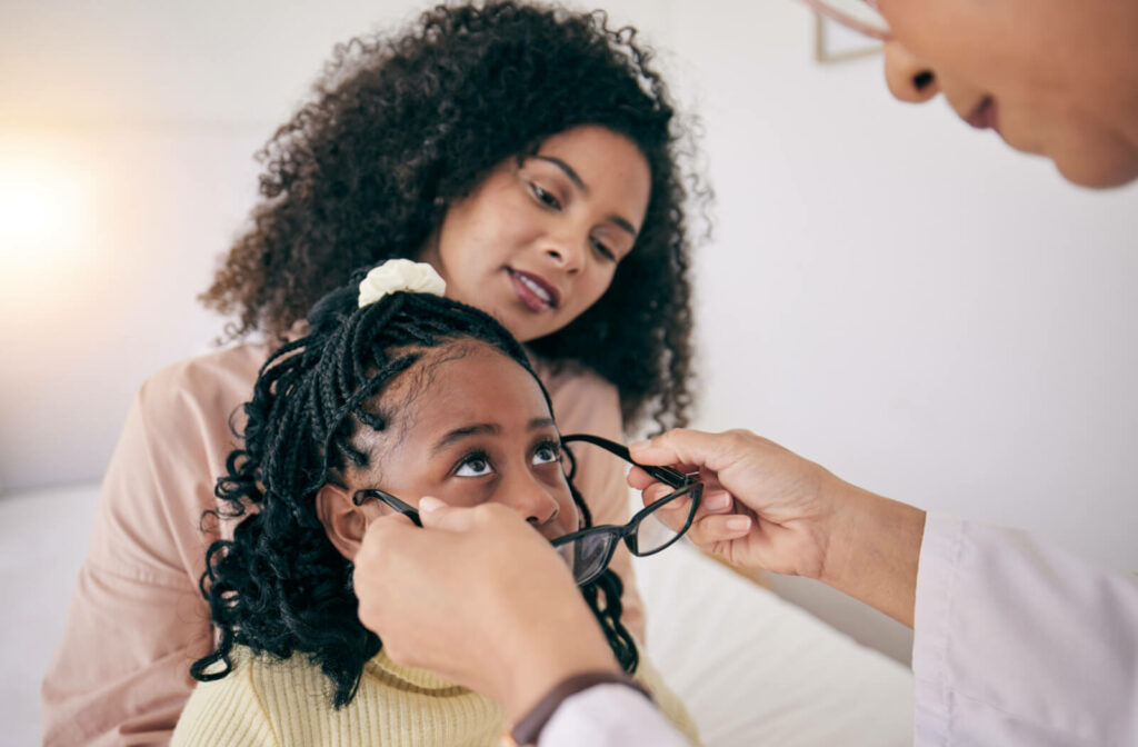 A child with her mother trying on glasses in a store while being assisted by an optician or optometrist.